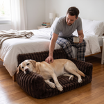 Man sitting on the edge of his bed with a morning coffee, scratching a yellow Labrador resting in the HuddleHaven dark brown dog sofa bed